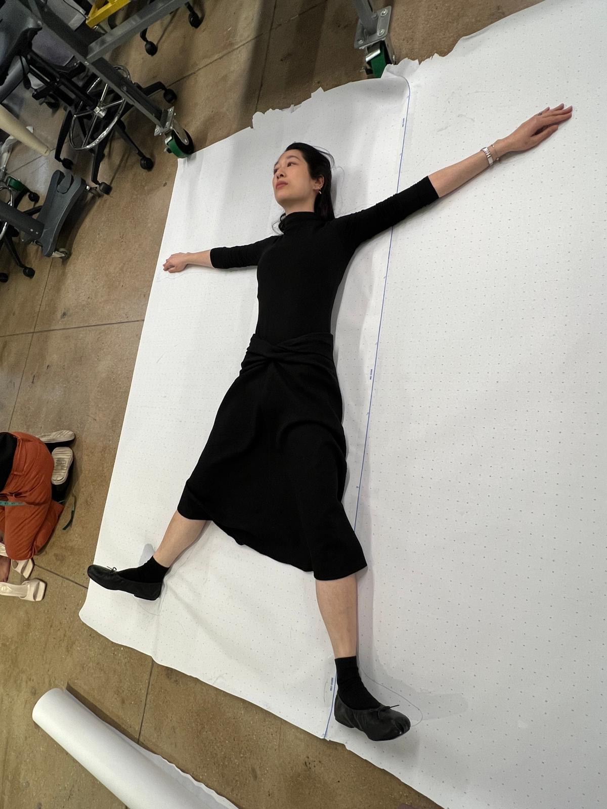 Deborah Won draping on herself during MFA, lying on pattern paper with arms extended